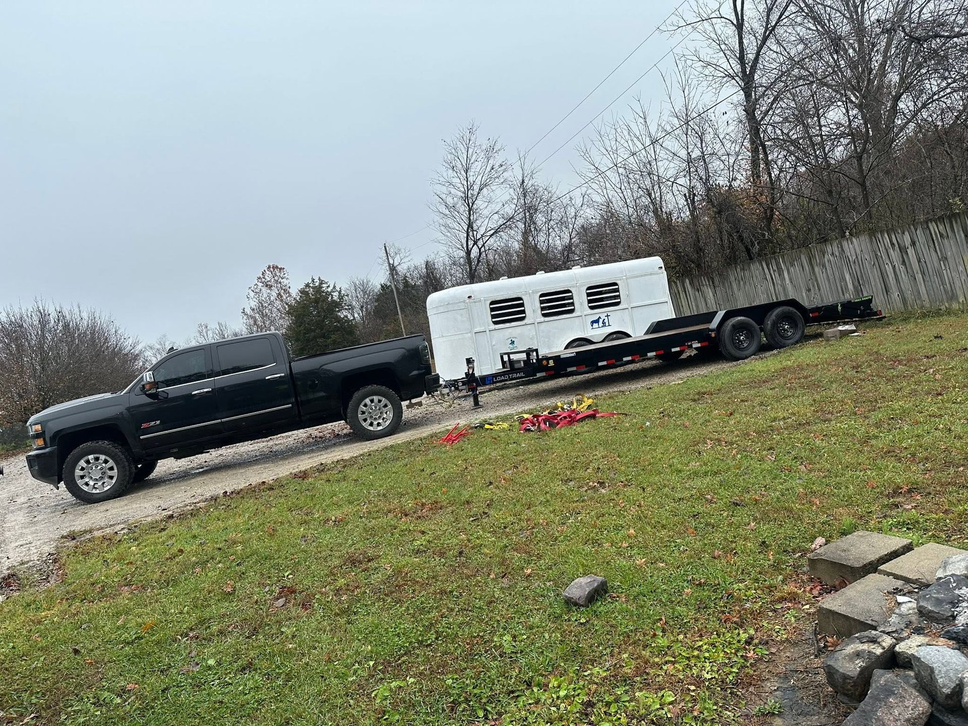 Black pickup truck towing a white trailer on a gravel hill beside a grassy yard and trees