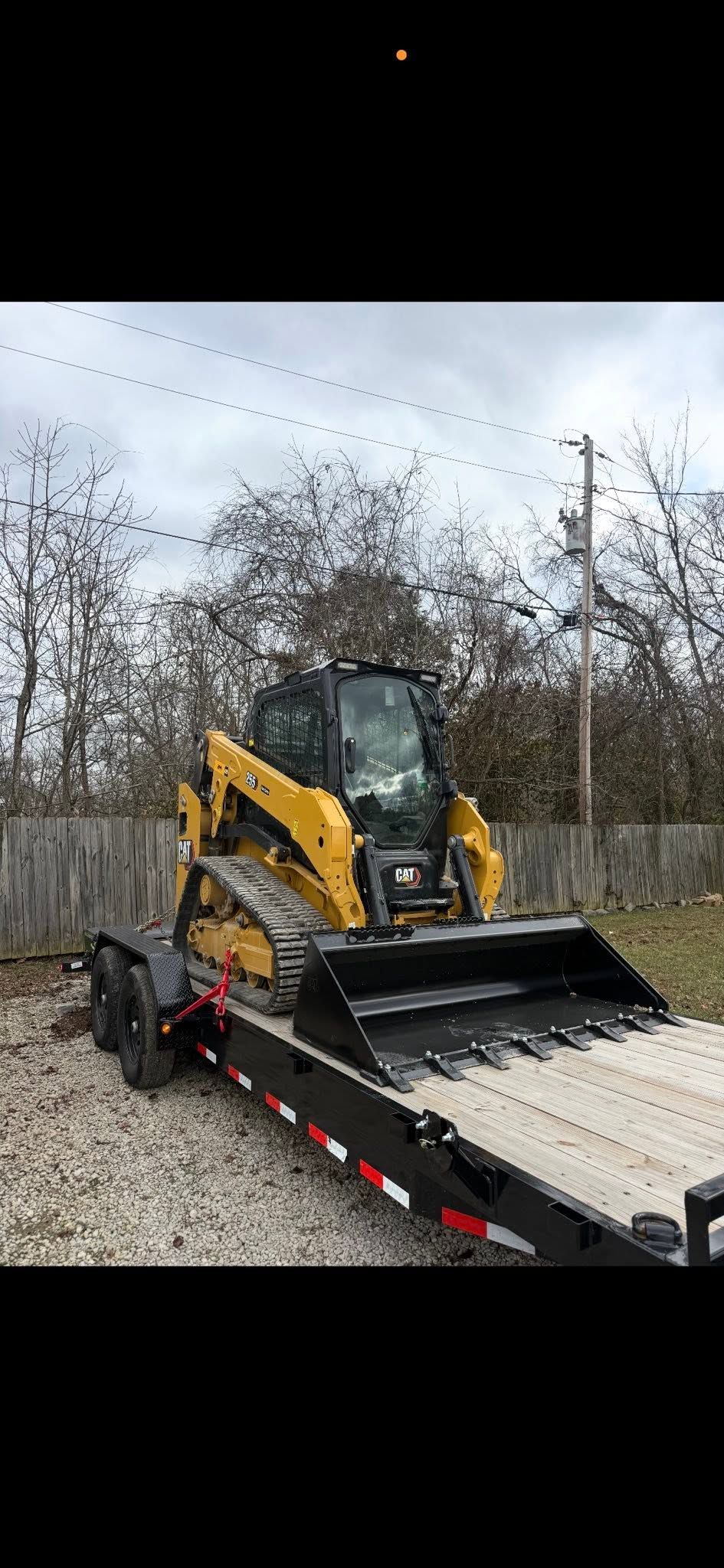 Yellow-and-black skid steer on a flatbed trailer in a gravel lot, with trees and an overcast sky behind it