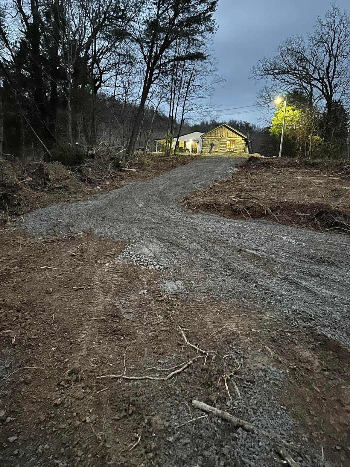 Gravel road at dusk leading to a lit yellow house beside bare trees