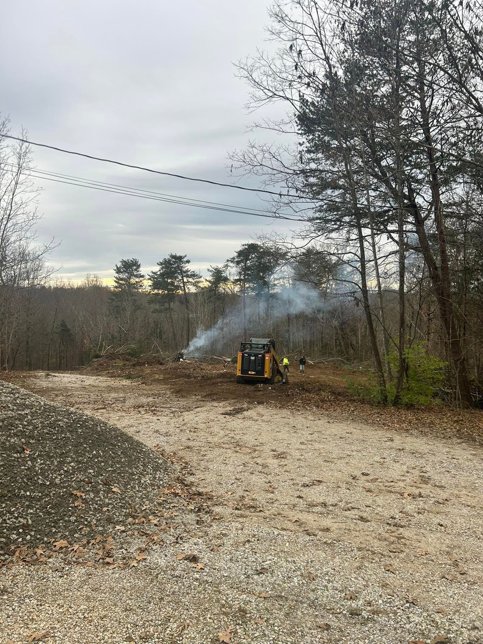 Smoking fire pit beside a gravel path in a wooded area under overcast sky