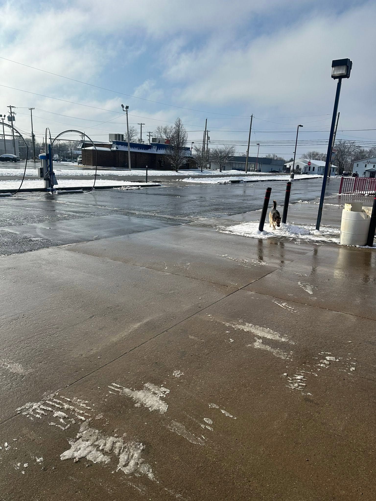 Snowy parking lot with wet pavement, streetlights, and distant traffic under a cloudy sky
