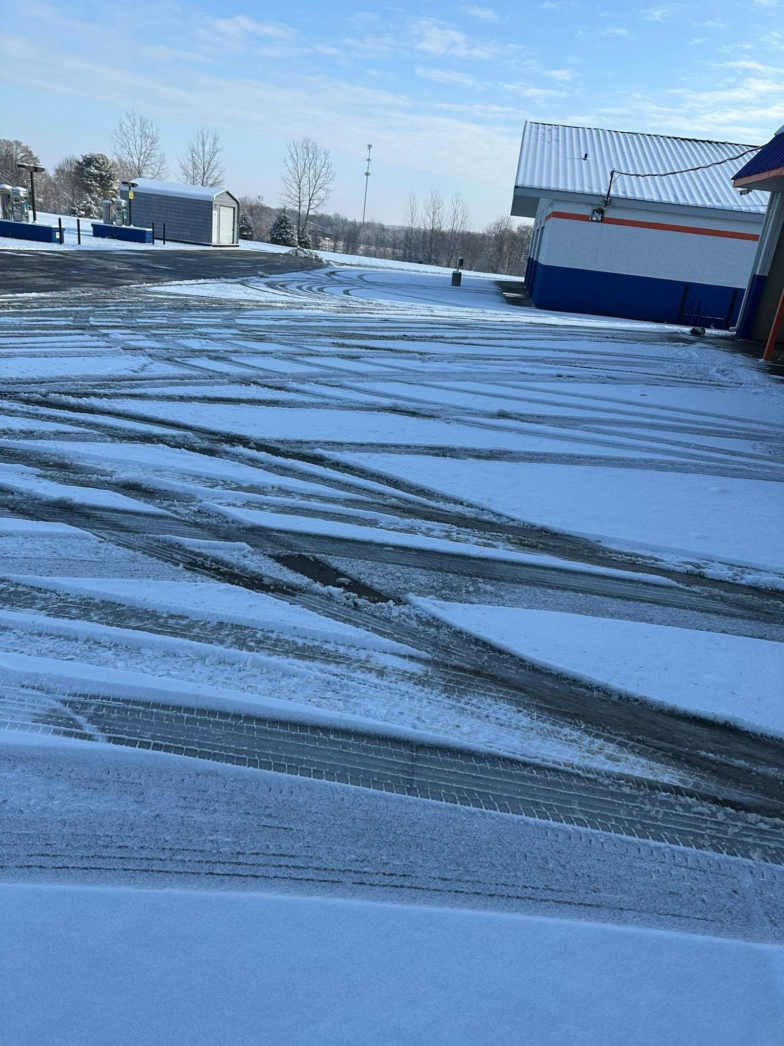 Snow-covered parking lot with tire tracks and a small building under a clear blue sky