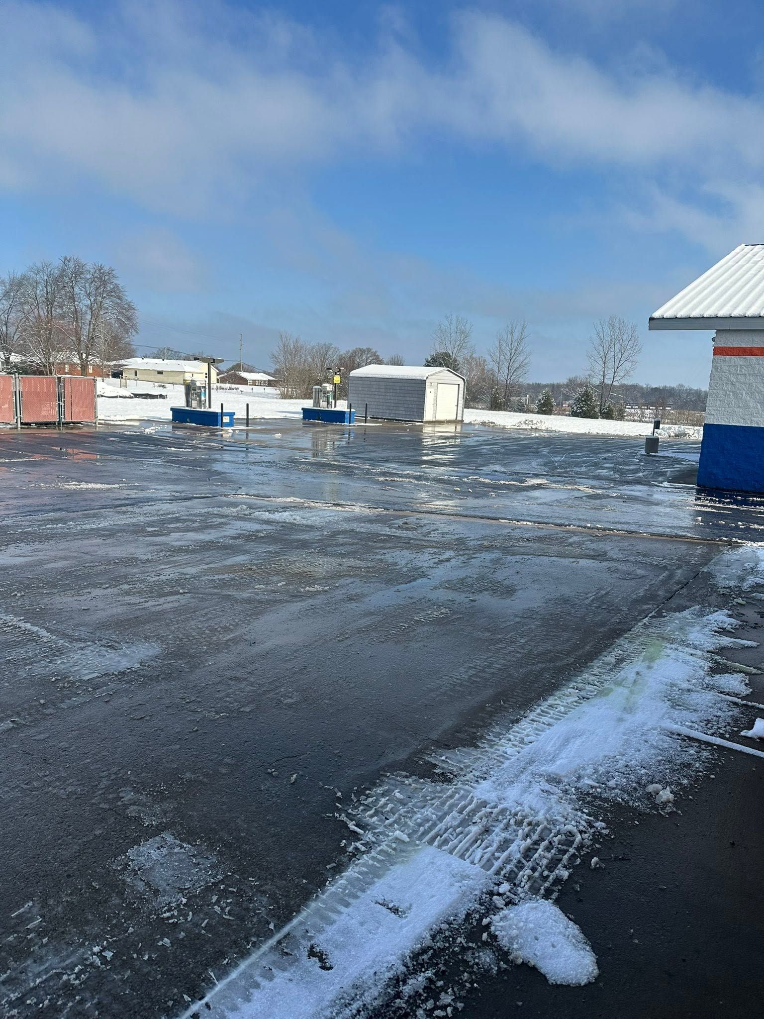 Snowy parking lot with patches of ice, small buildings, and a clear blue sky