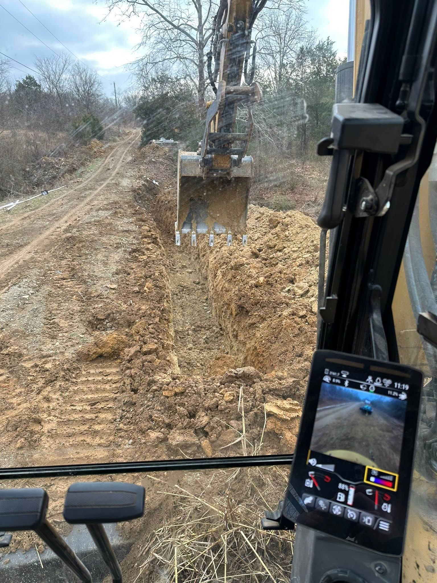 Excavator bucket clearing a leaf-covered dirt path in a wooded area, seen from the cab.