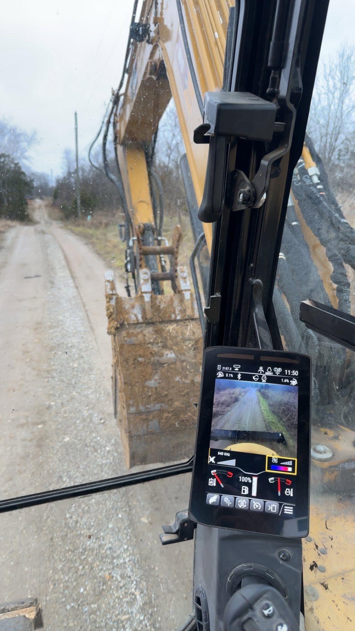 View from inside an excavator cab on a dirt road, with the excavator arm visible outside.