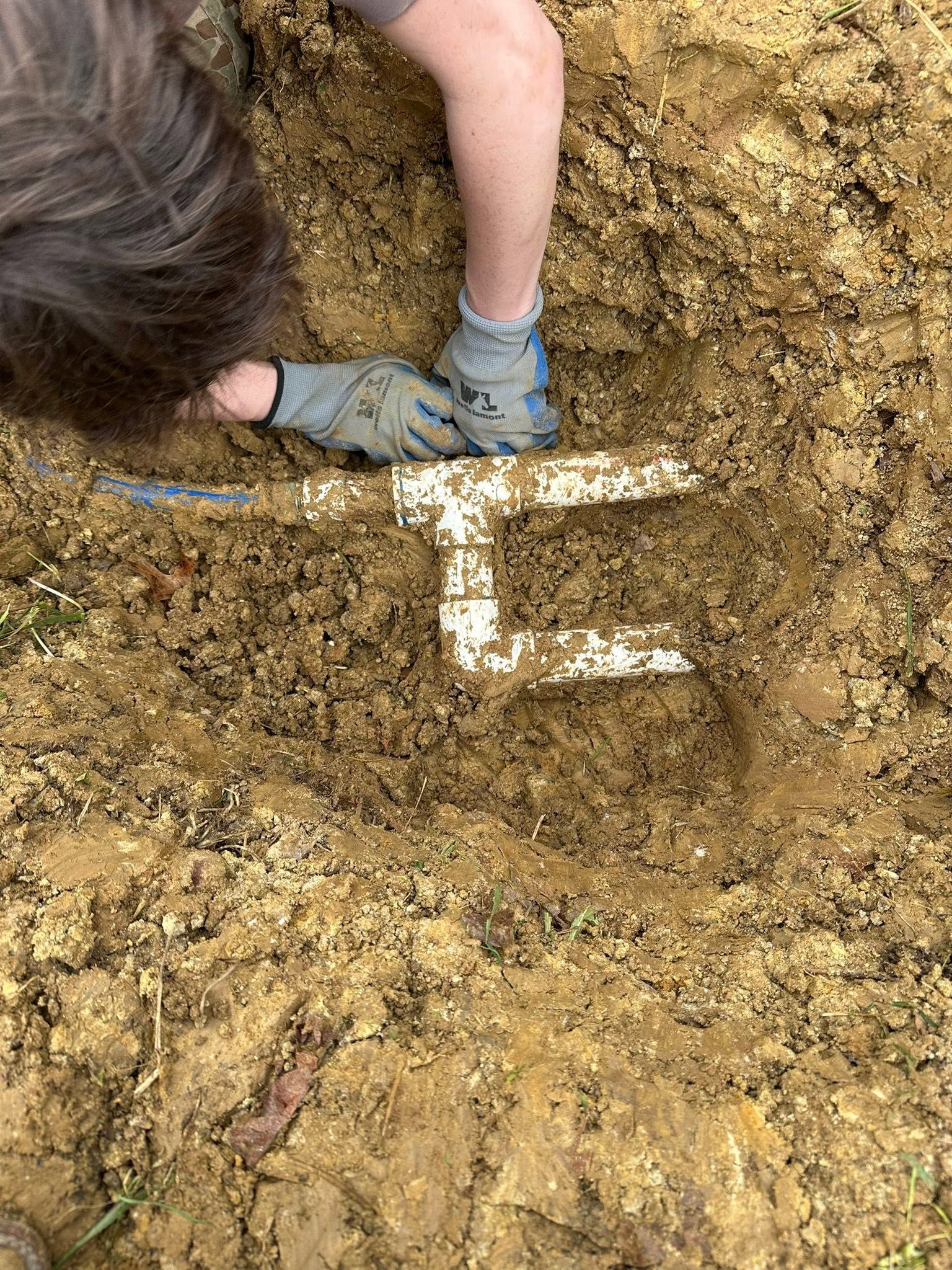 Person kneeling in muddy soil beside a white marker or measuring tool.