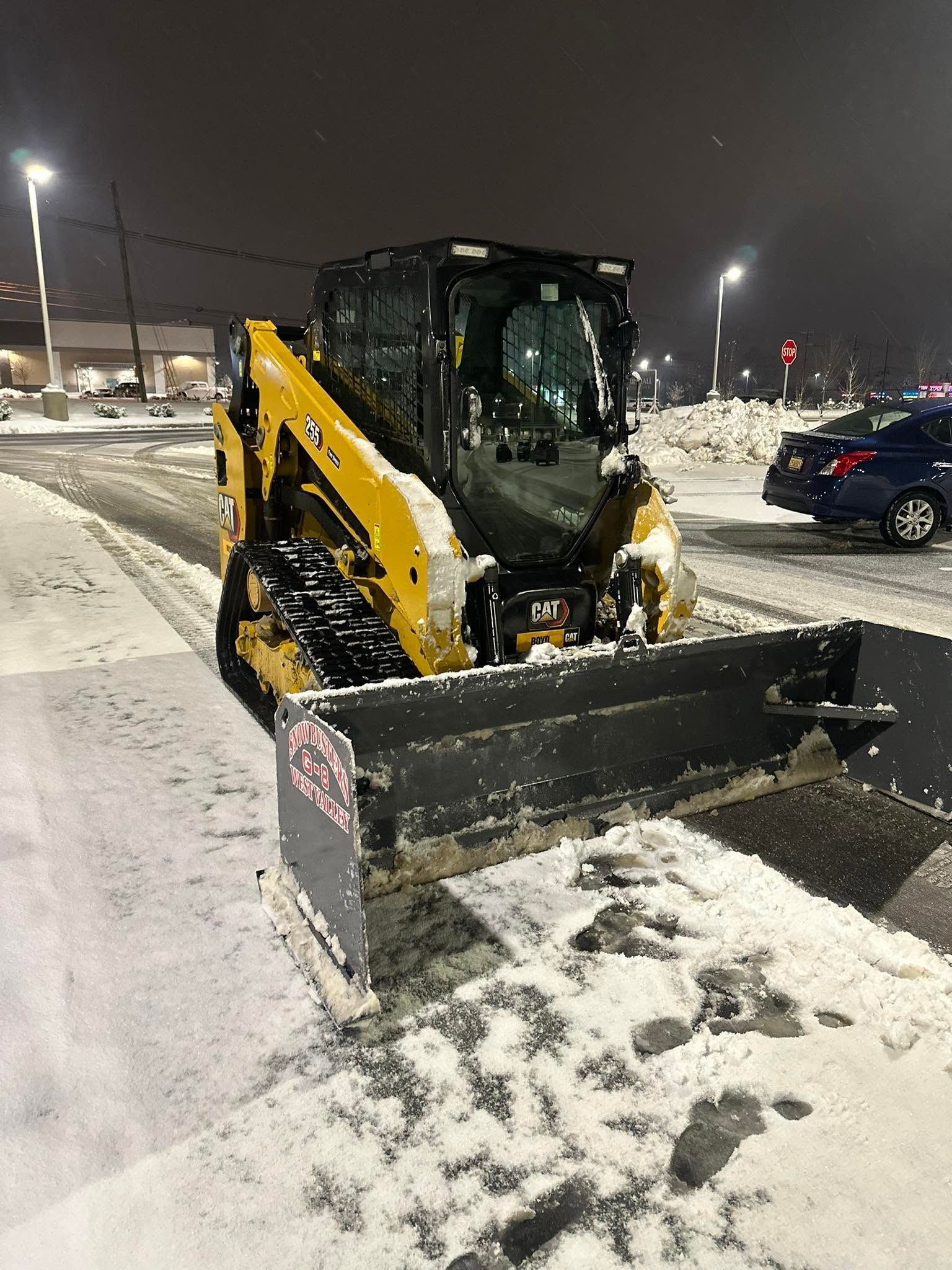 Yellow skid steer with front plow clearing snow in a parking lot at night