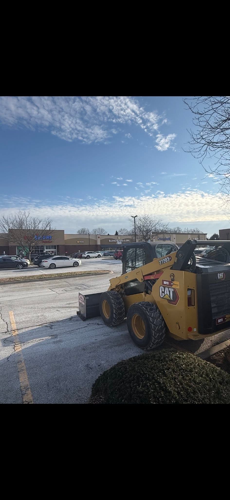 Yellow construction loader on a snowy lot under a blue sky, with parked vehicles and buildings in the background