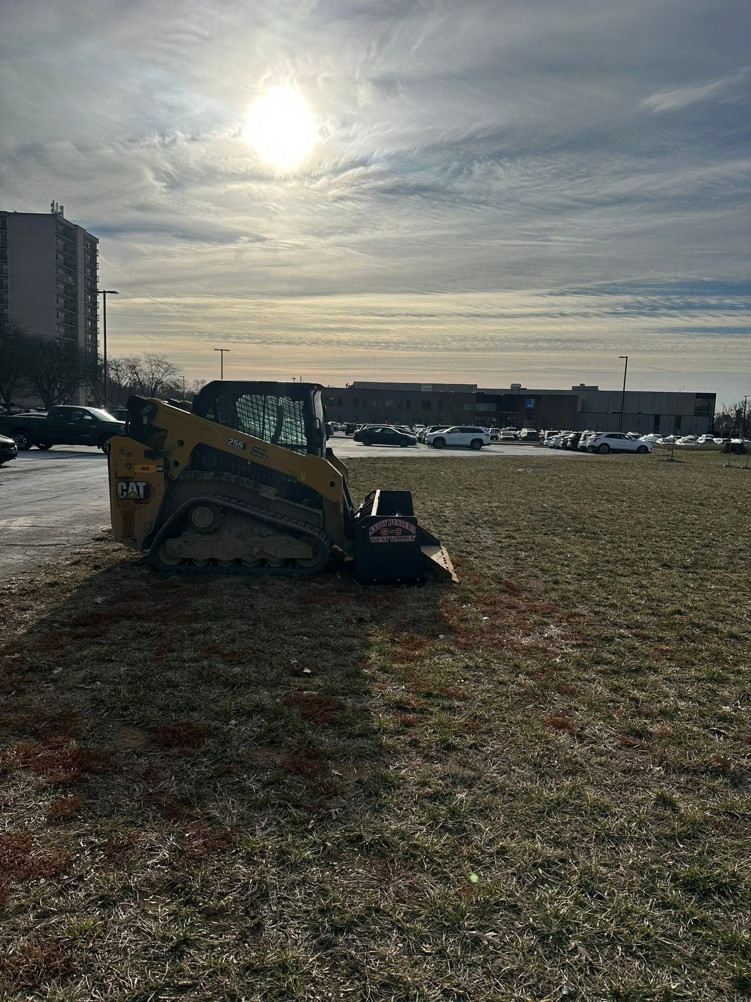 Yellow excavator on a grassy field under a bright, cloudy sky near the waterfront