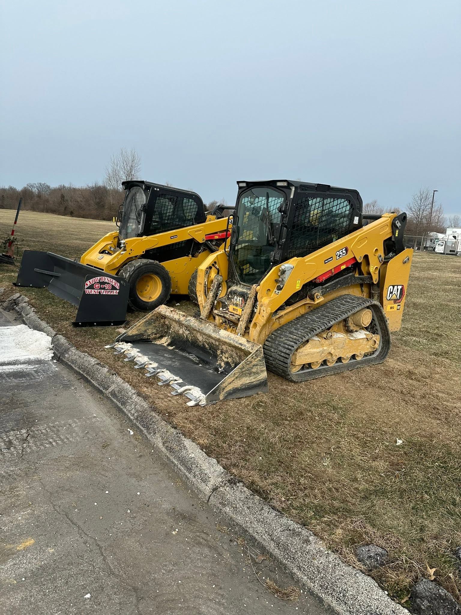 Two yellow skid steer loaders parked on a gravel edge beside a road under a cloudy sky