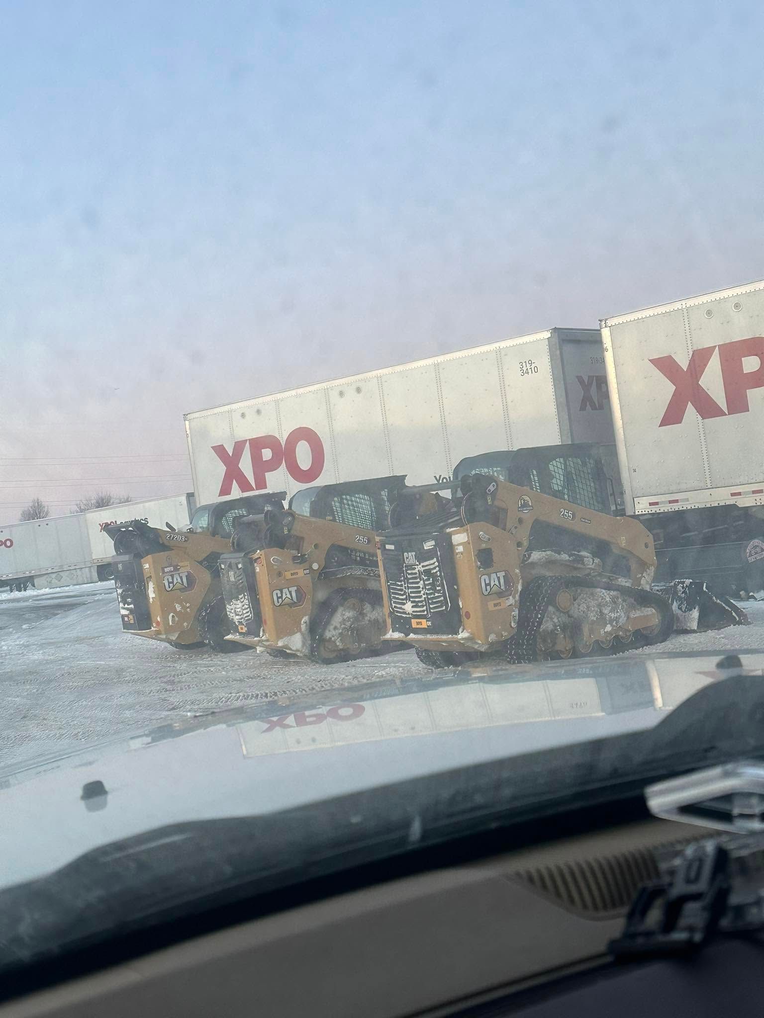 Yellow bulldozer loading debris by an XPO truck on a snowy roadside, seen from inside a car