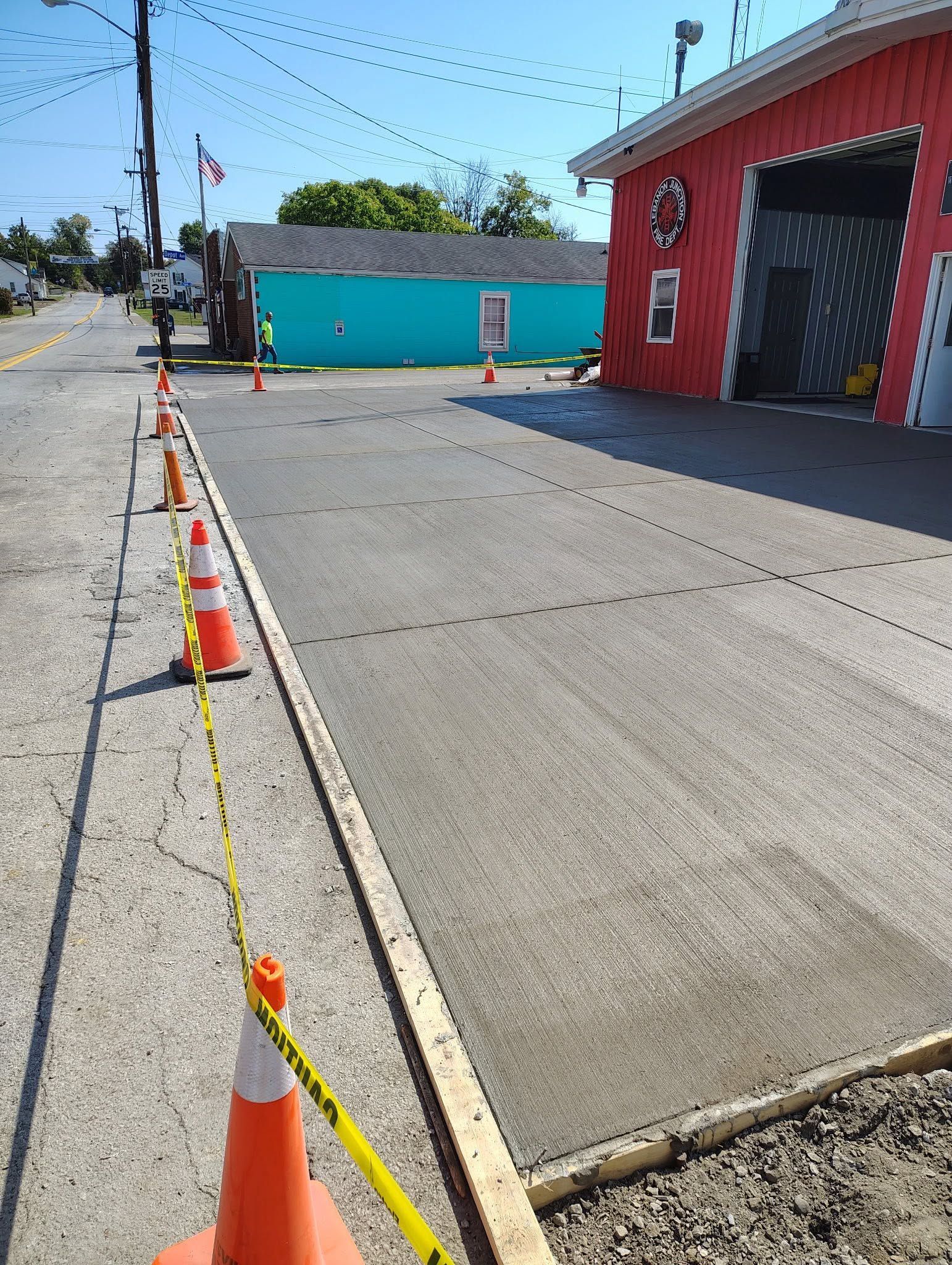 Orange cones and yellow tape line a concrete walkway beside a red building and blue wall on a sunny street.