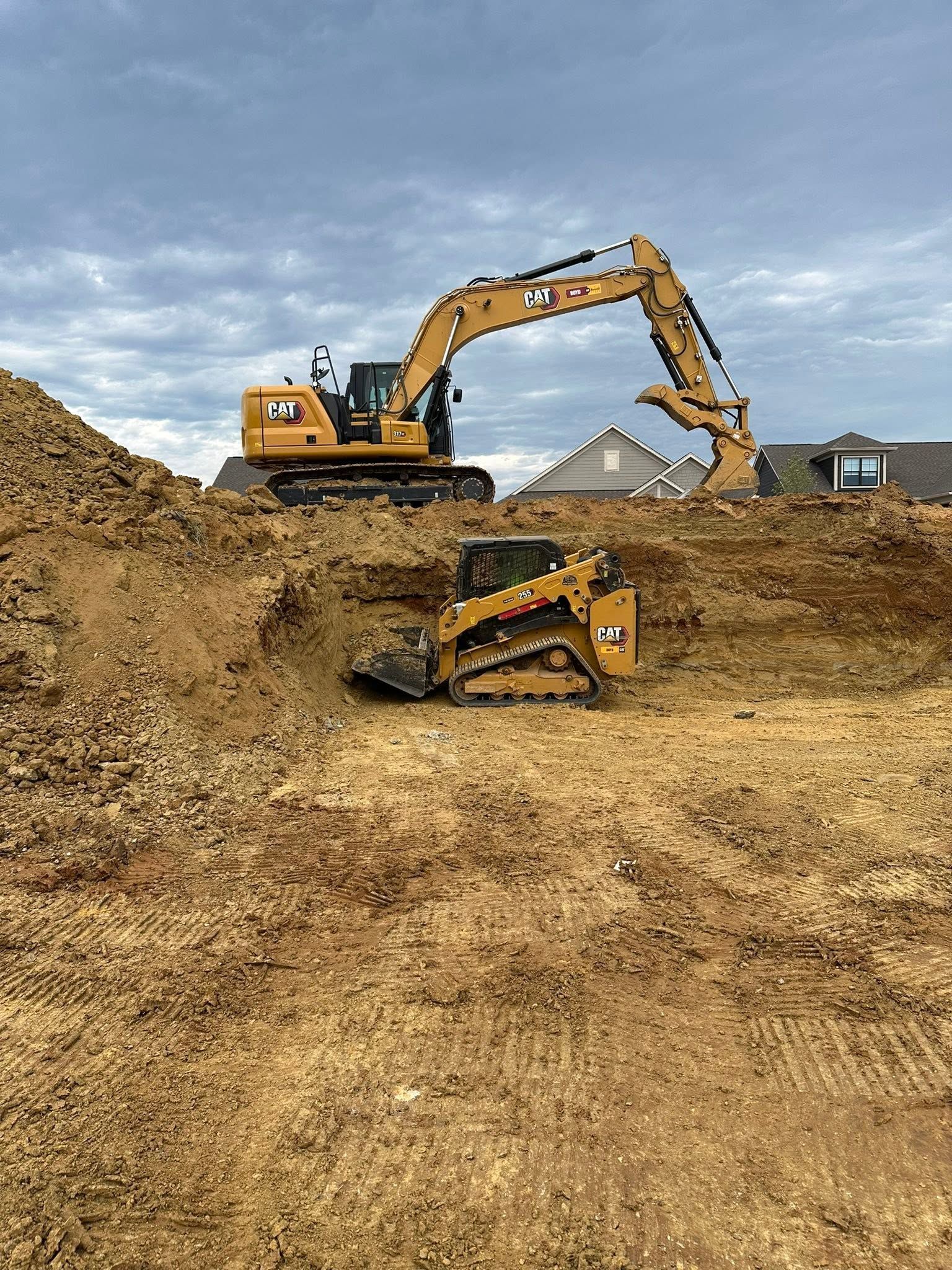 Yellow excavator working on a dirt mound under a cloudy sky, with a tipped loader in the foreground.