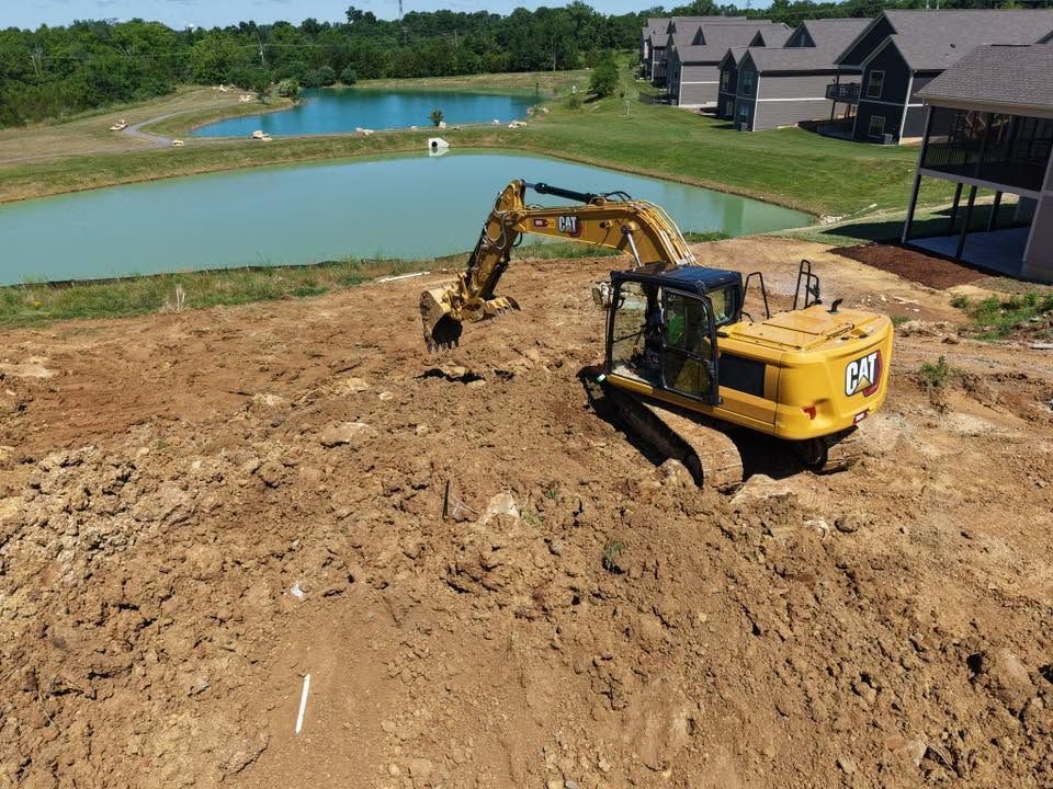 Excavator and dump truck grading a dirt lot beside a pond and new houses