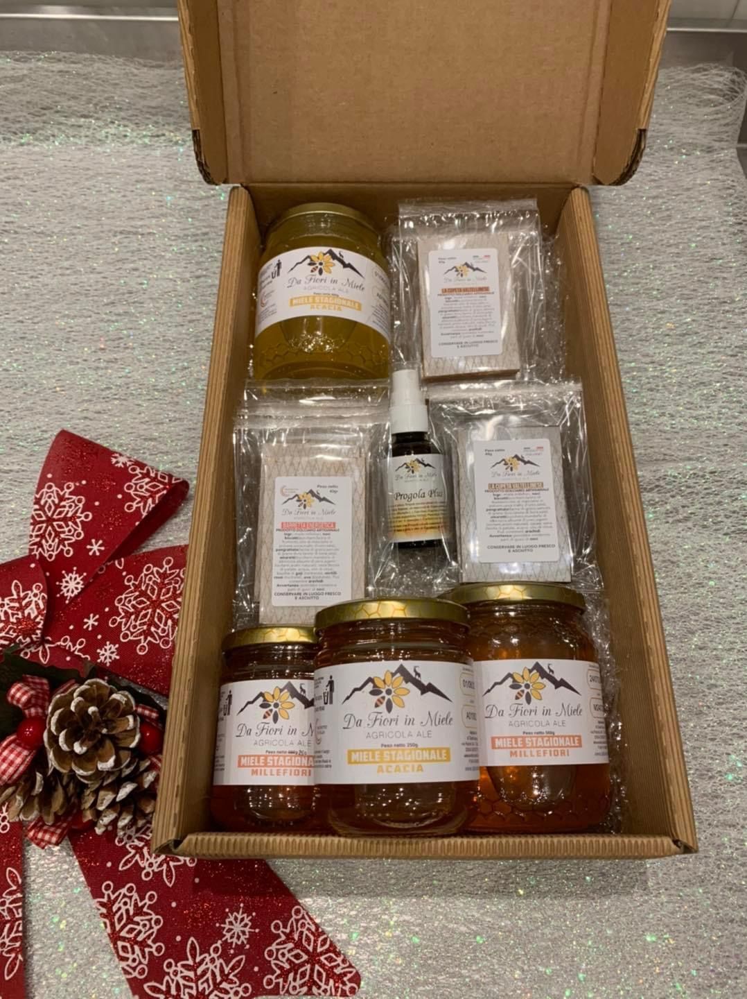 Scatola regalo piena di barattoli di miele, bustine e flacone contagocce, accanto a un fiocco festivo.