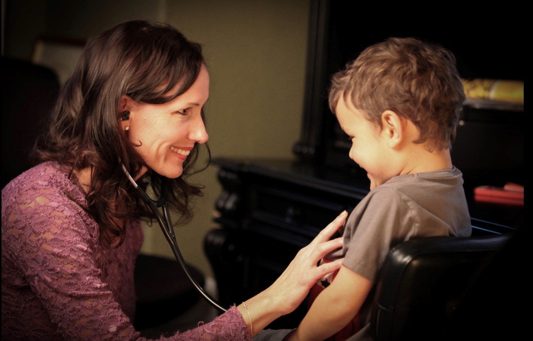 Doctor listening to a young boy's chest with a stethoscope. Both are smiling in a dimly lit room.