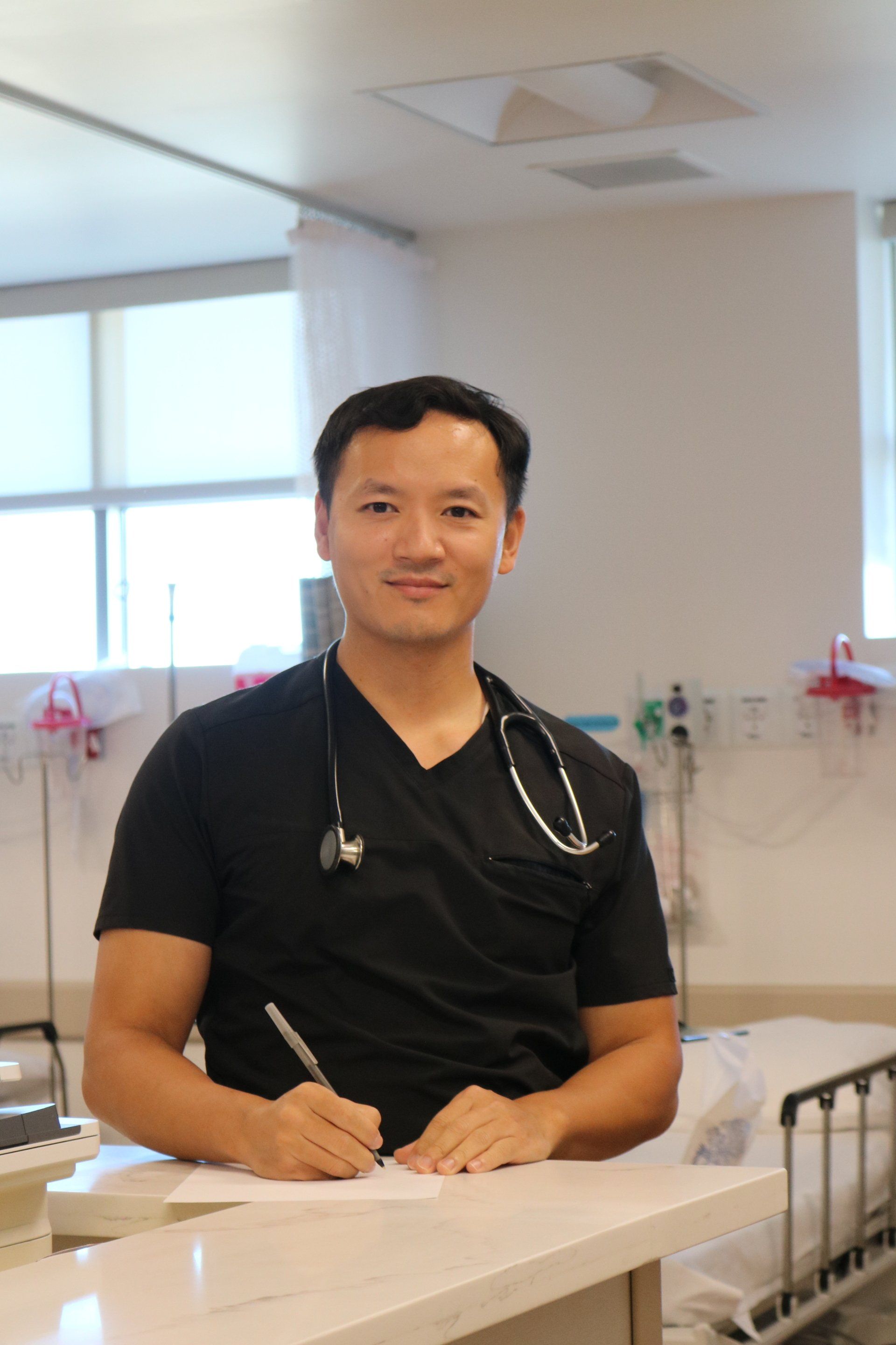 Asian doctor in scrubs writing at a counter in a hospital room, stethoscope around his neck.