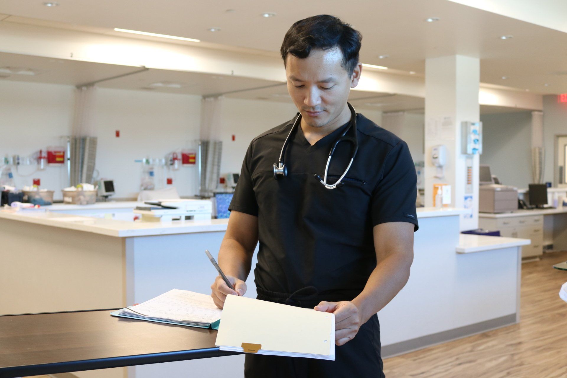 Doctor in black scrubs writes on a chart in a medical setting with patient bays visible.