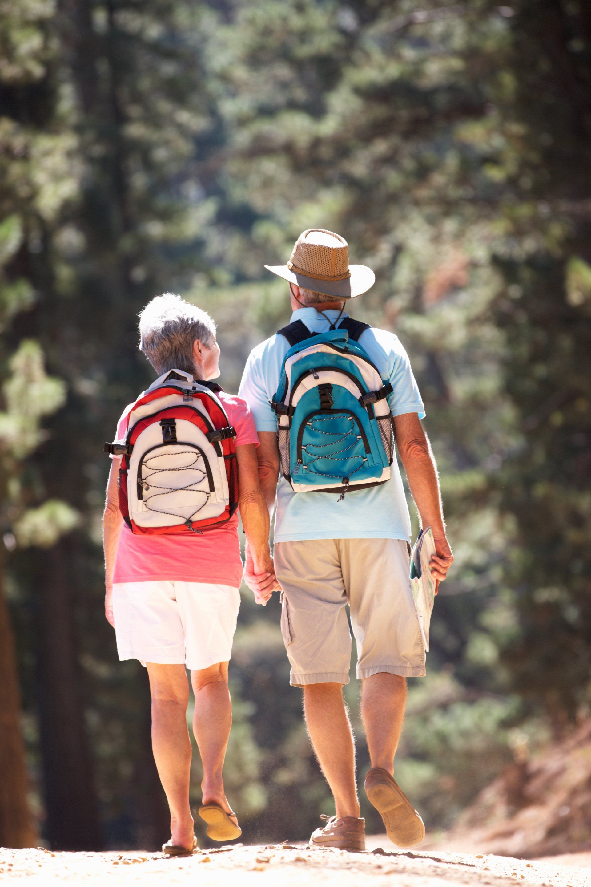 Elderly couple hiking, holding hands, with backpacks on a wooded trail.