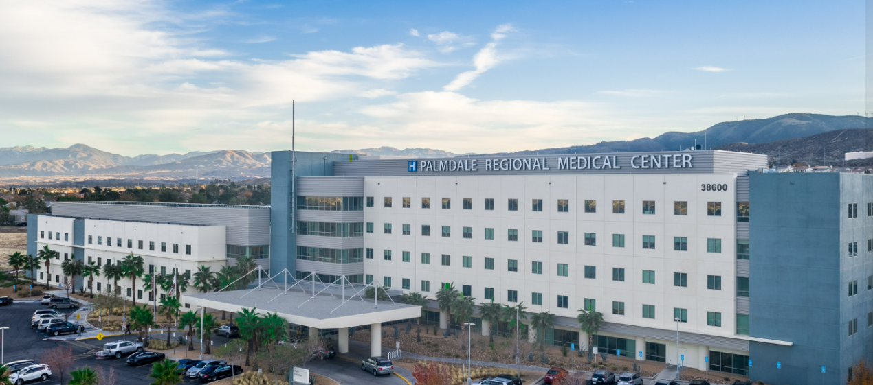 Exterior view of Victor Valley Global Medical Center. White building with blue accents, palm trees, and mountain backdrop.