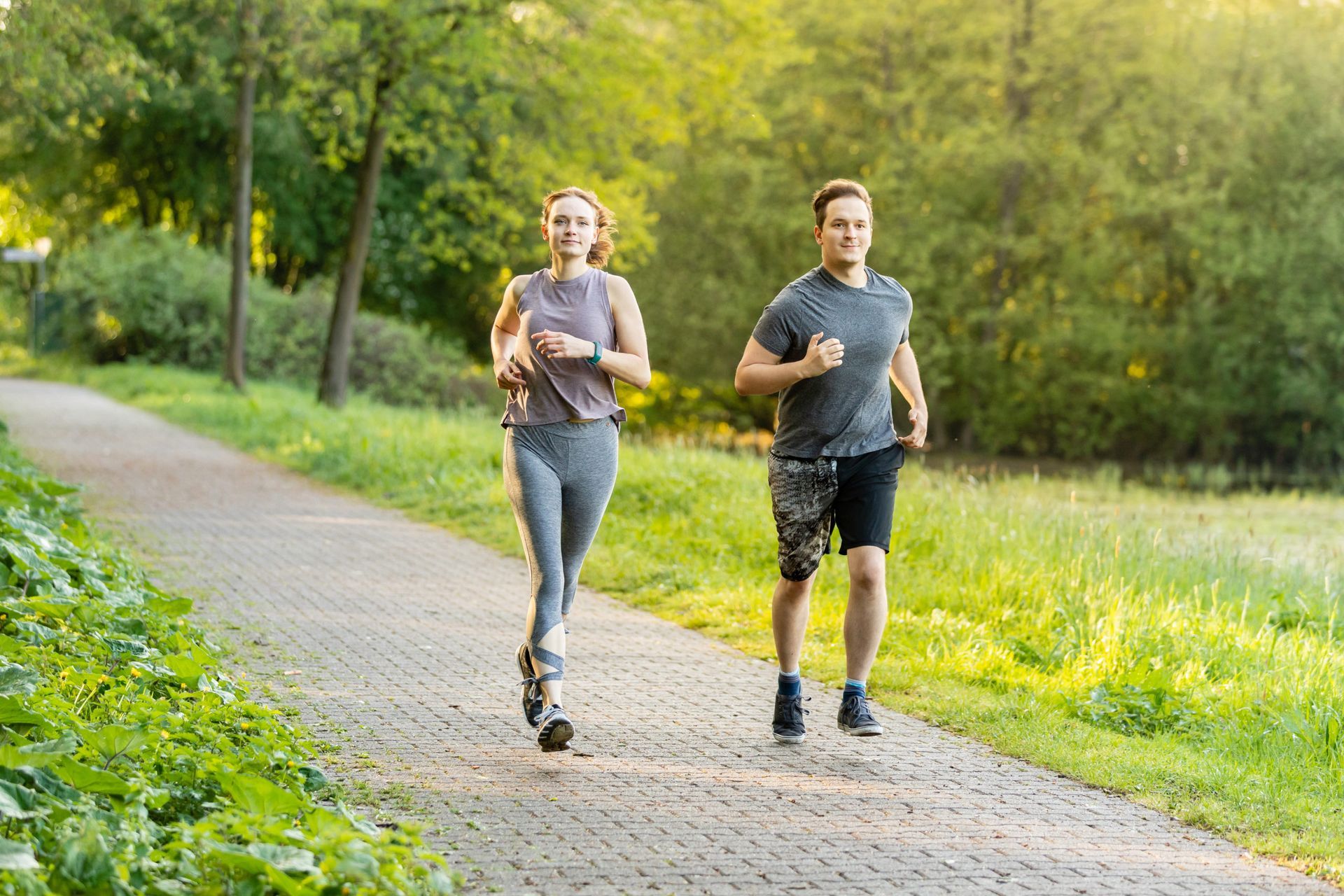 Woman and man jogging on a paved path in a park; sunny day, green foliage.