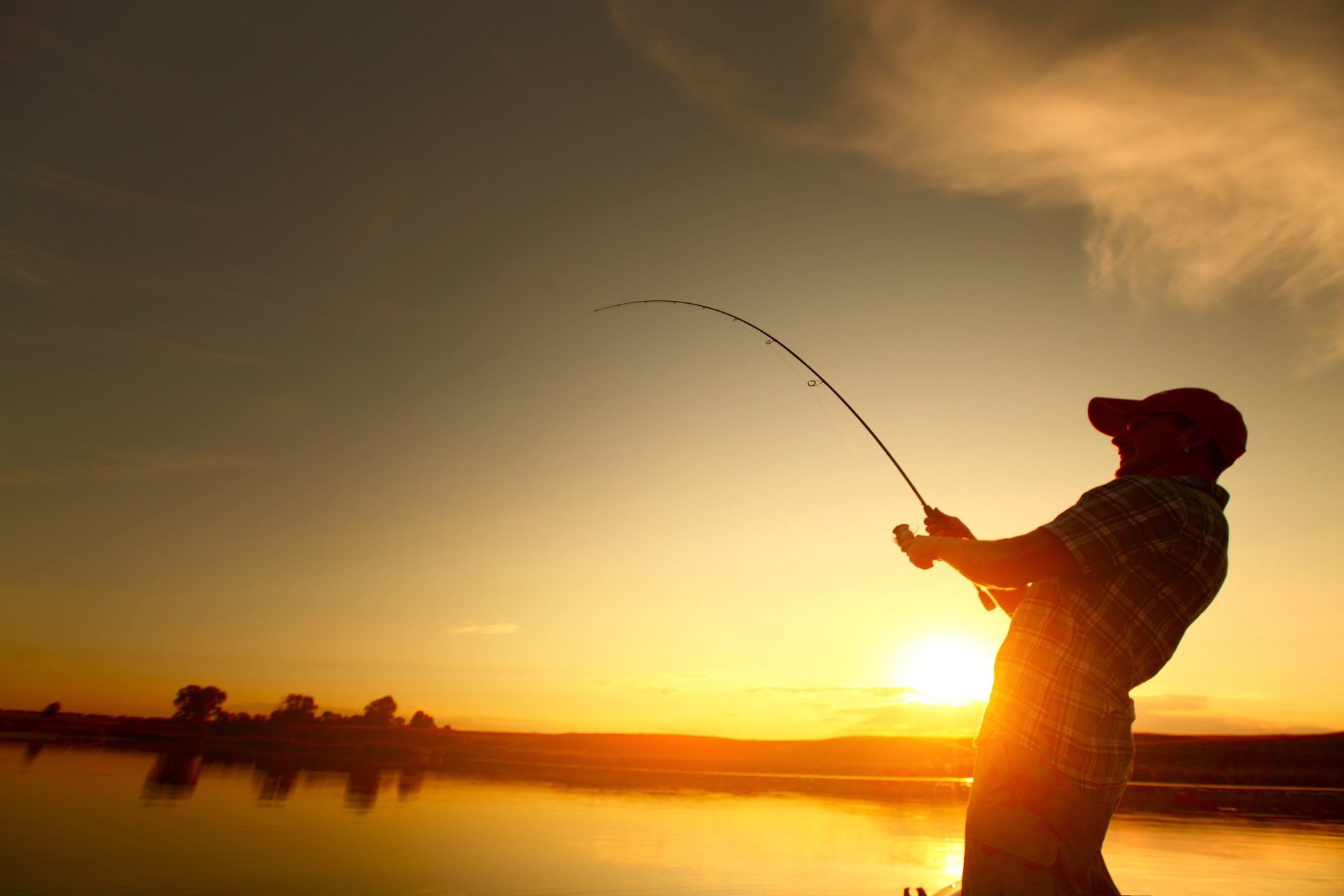 Silhouette of person fishing at sunset, rod bent, casting line over water.