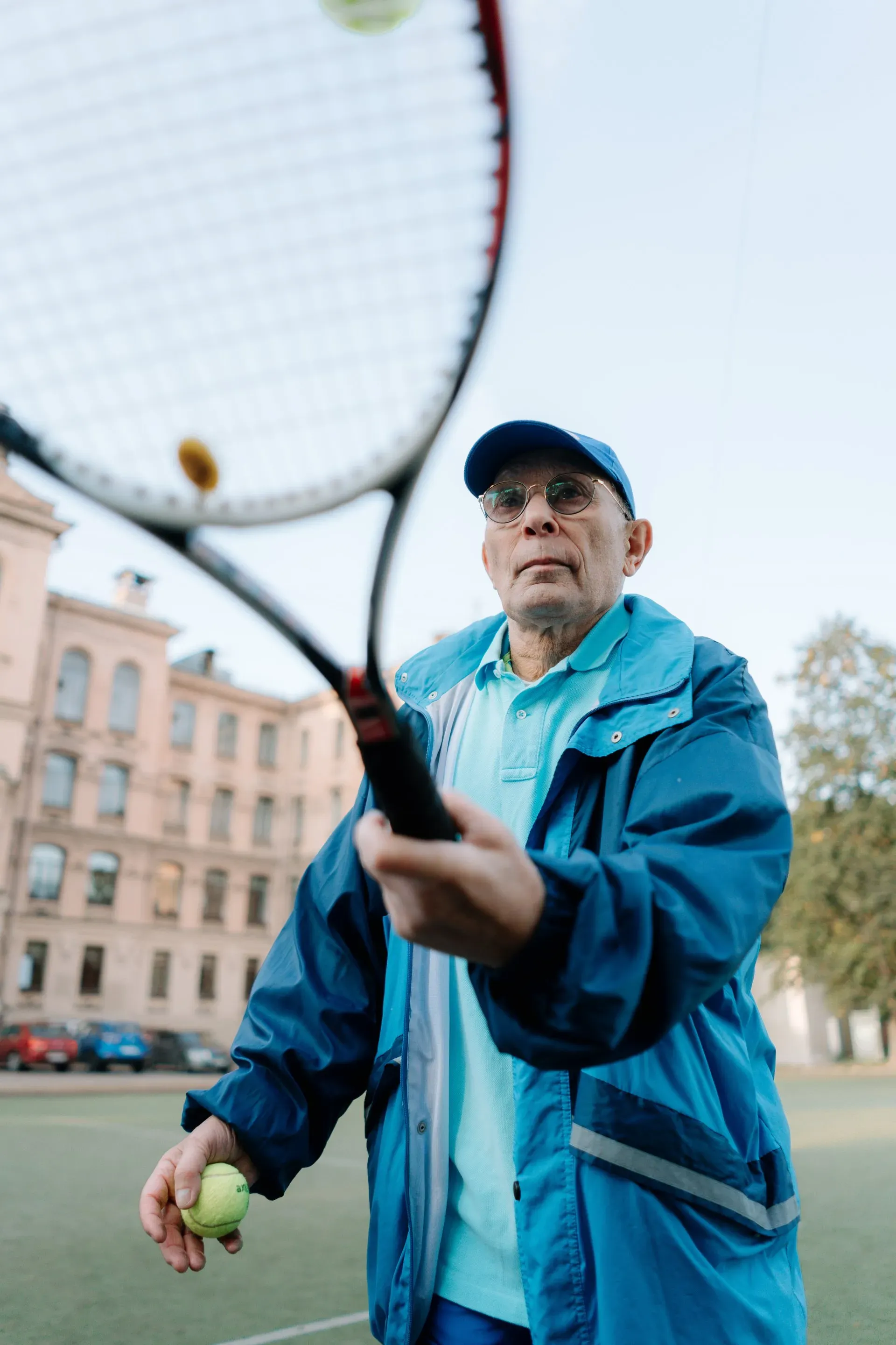 Man in blue jacket and cap prepares to serve a tennis ball, holding racket, outdoors.