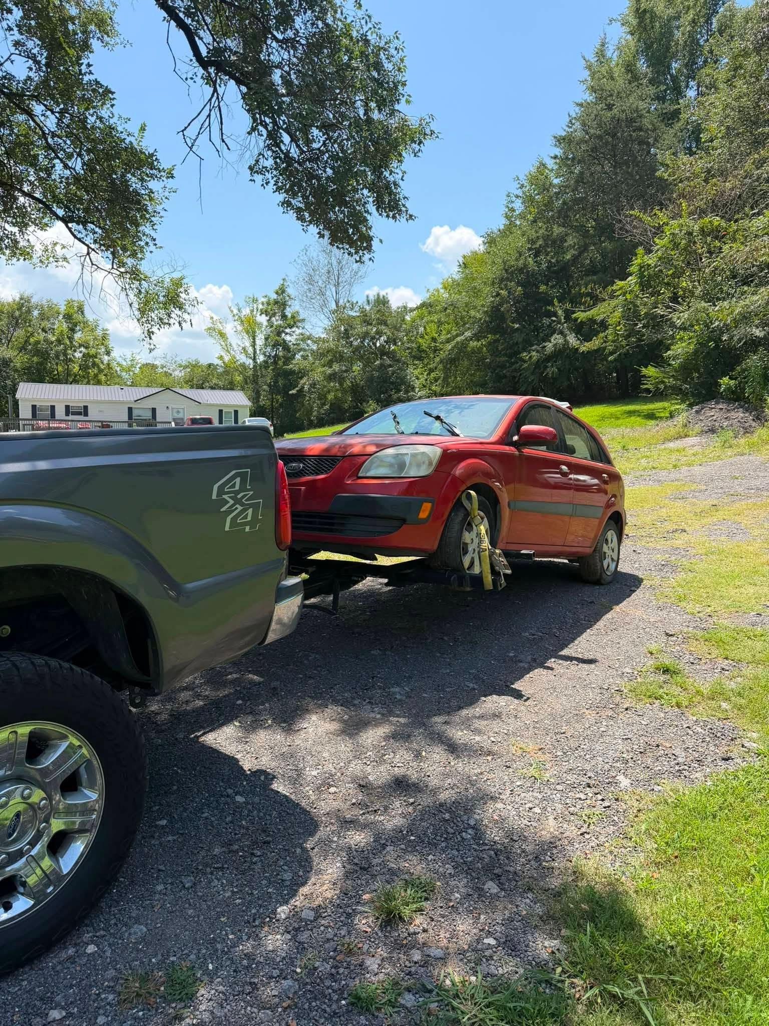 Red car parked nose-to-nose with a dark green truck on a gravel driveway under trees.