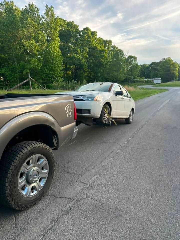 White sedan with rear-end damage hitched to a tow truck on a roadside near trees.