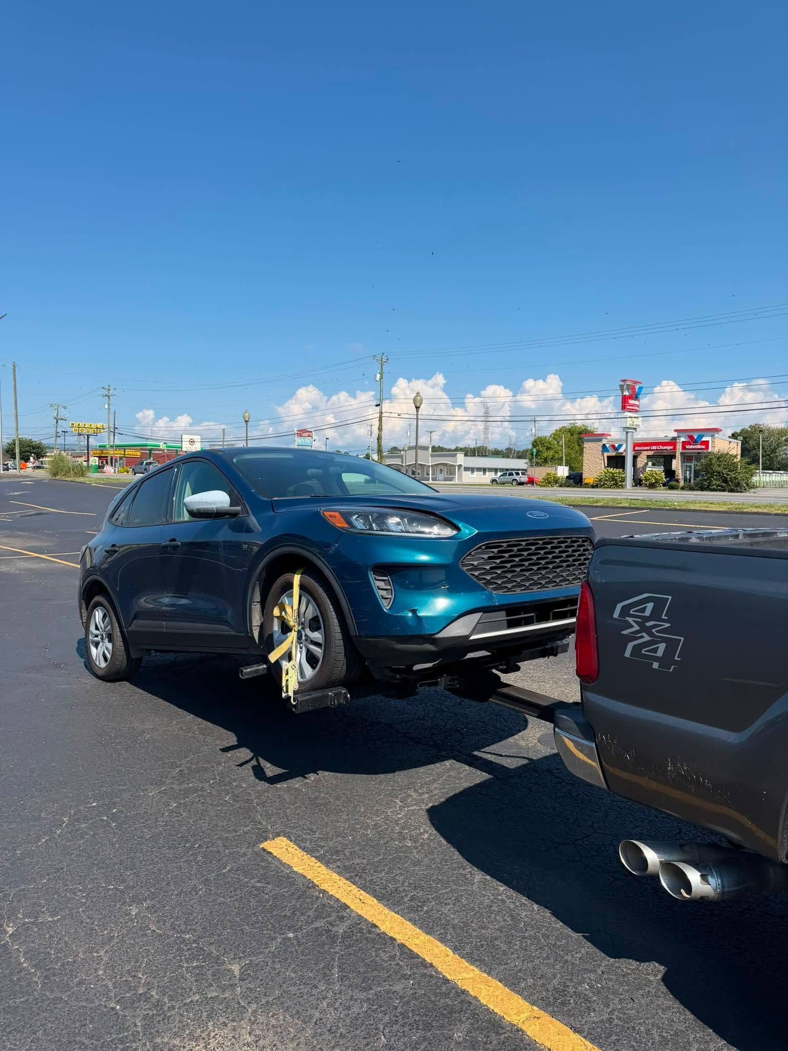 Blue car towing a black trailer on a sunny road, with one wheel lifted off the ground.