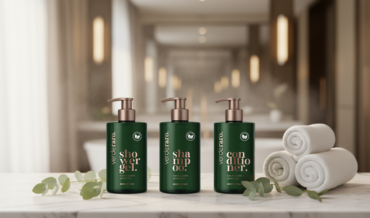 Tre flaconi con dosatore di colore verde scuro, etichettati rispettivamente bagnoschiuma, shampoo e balsamo, sono appoggiati accanto ad asciugamani bianchi su un ripiano di marmo.