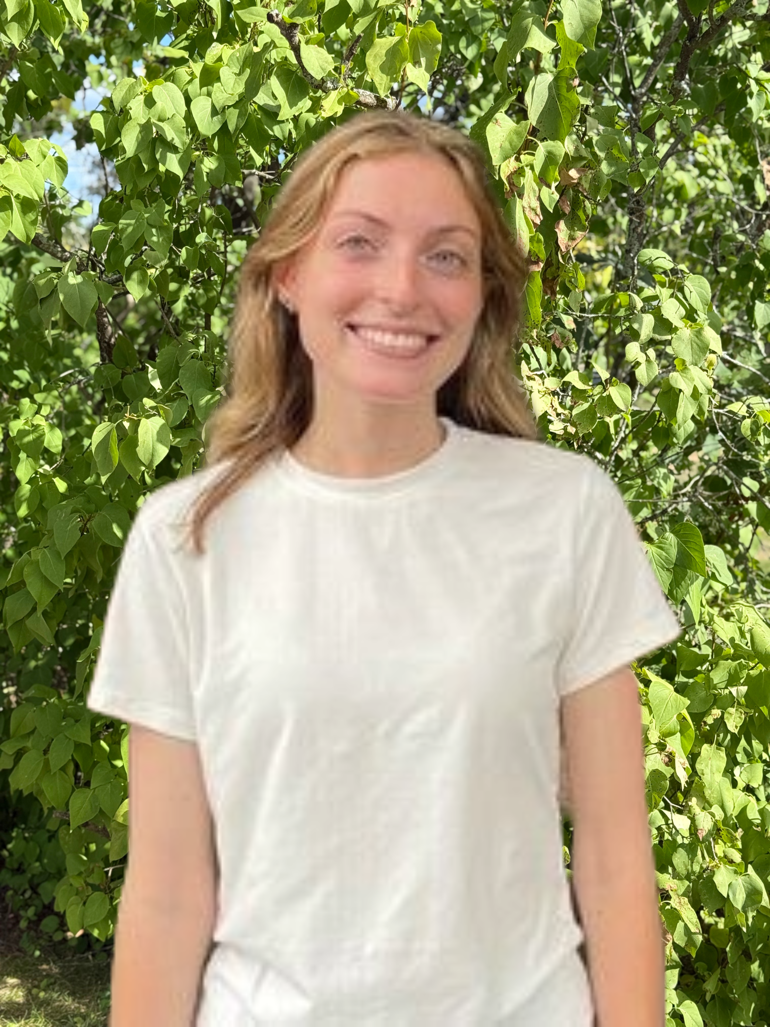 A woman in a white shirt is smiling in front of a tree.