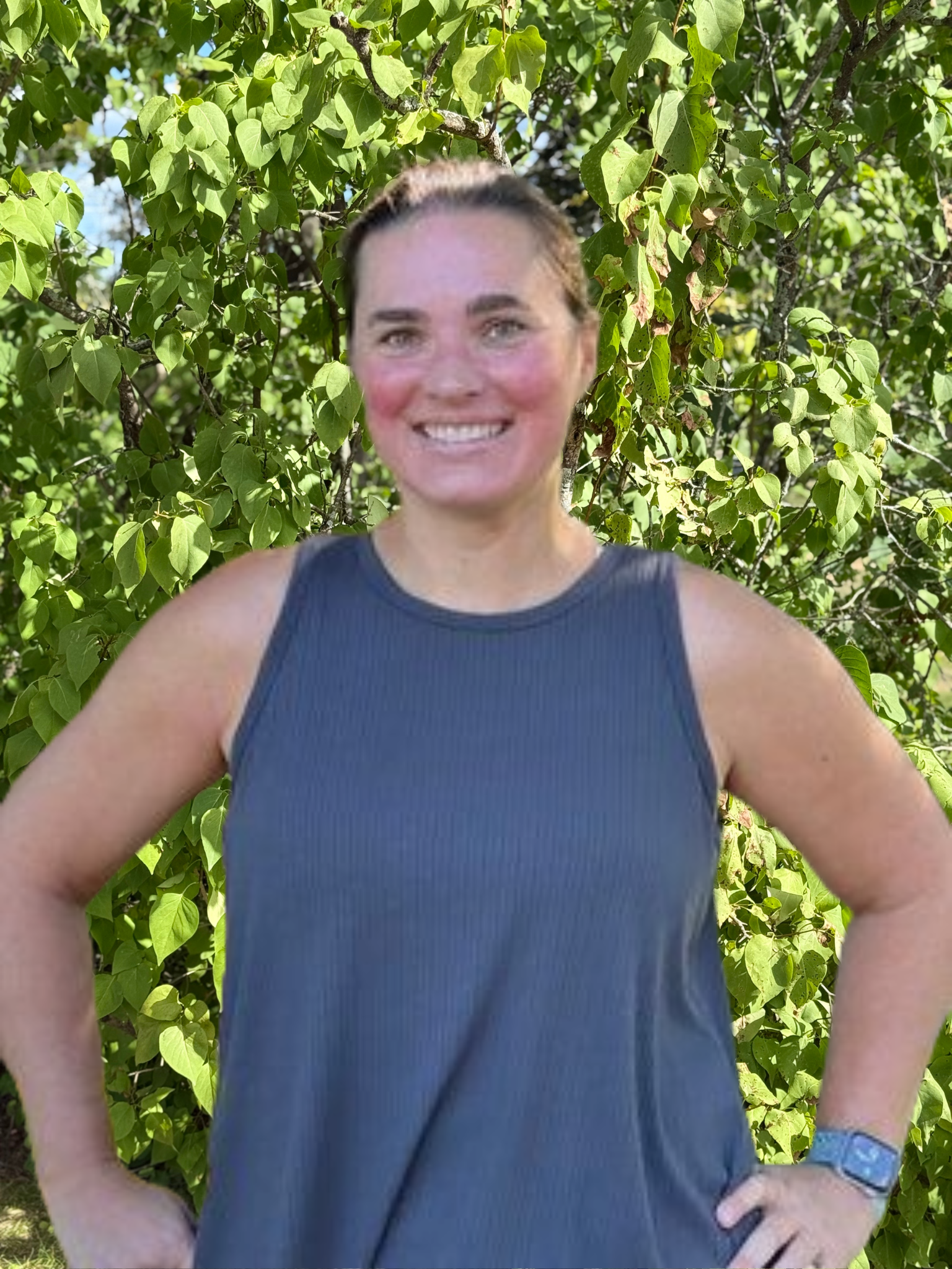 A woman in a gray tank top is standing in front of a tree with her hands on her hips.