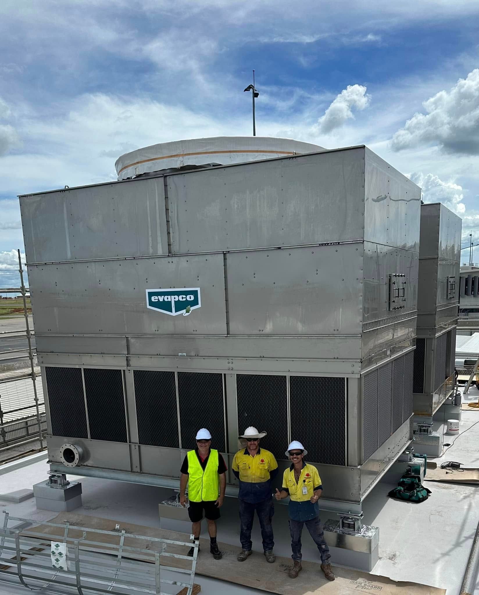 Three workers stand by a large metal cooling tower on a rooftop. Blue sky — Great Northern Airconditioning & Mechanical Services in Berrimah, NT