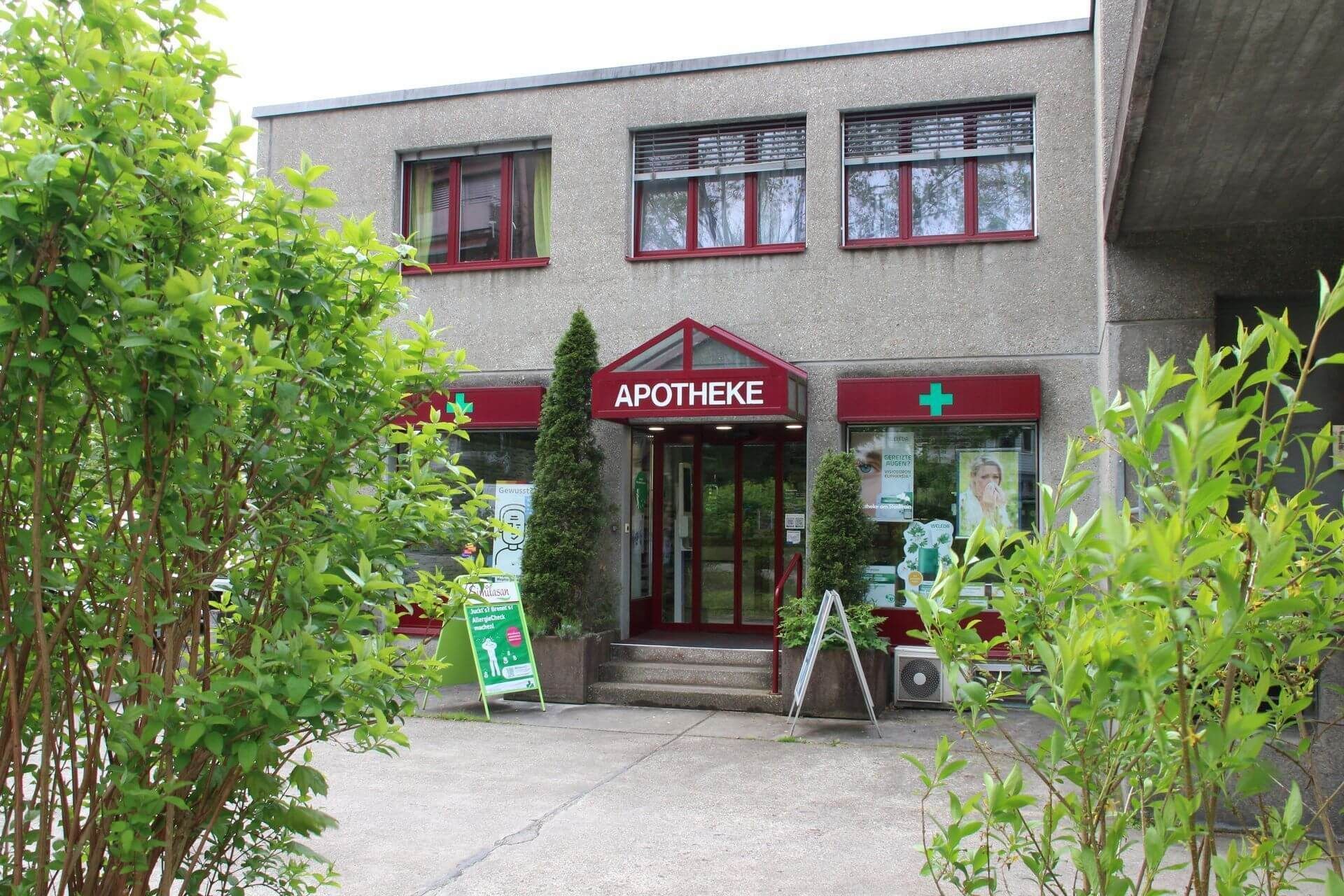 Apothekengebäude mit roten Akzenten, grünen Kreuzschildern und dem Schriftzug „APOTHEKE“ über dem Eingang.