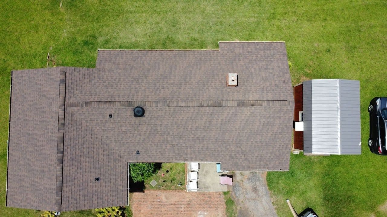 Aerial view of a large brown-roofed house with a patio, driveway, and detached metal garage on green lawn