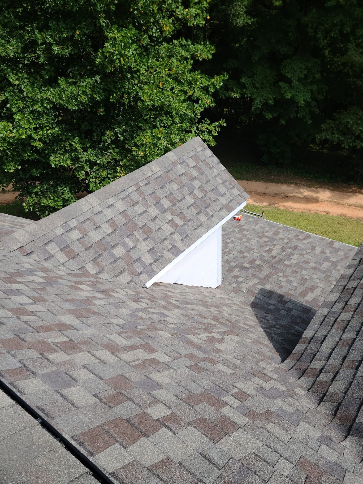 Rooftop with gray shingles and a white dormer, bordered by green trees.