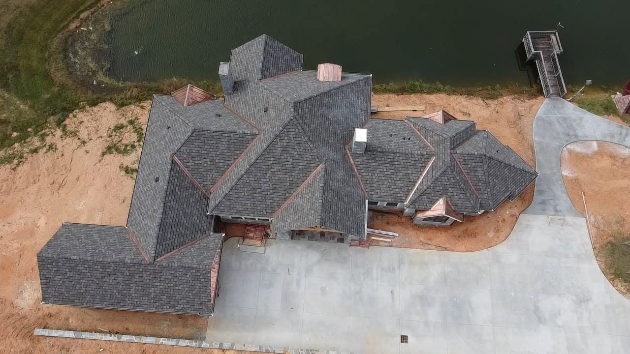 Aerial view of a dark-shingled house beside a driveway and lake, with a dock extending into the water.