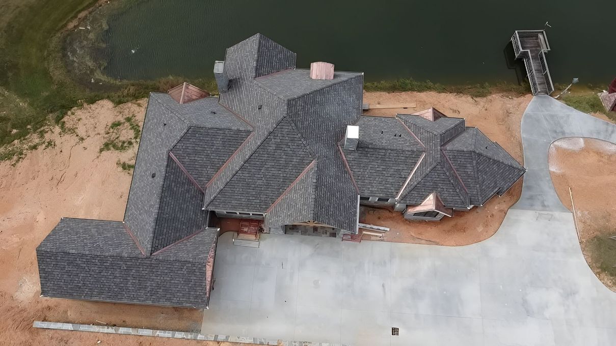 Aerial view of a large gray-roofed house beside water, with a driveway and dock nearby.