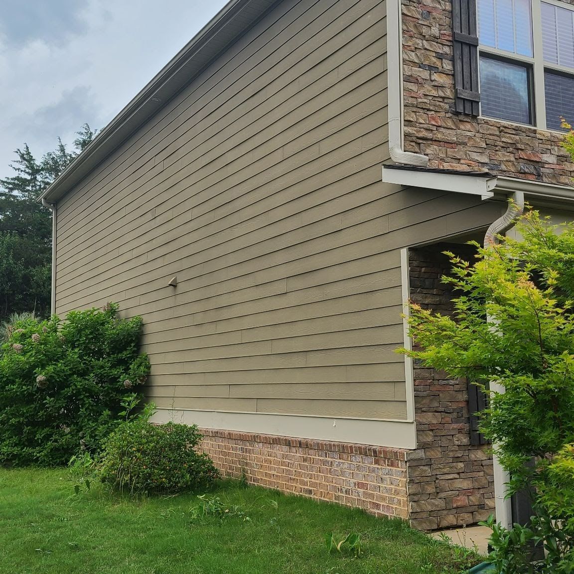 Side of a beige house with stone trim, white window frame, and green shrubs by the lawn