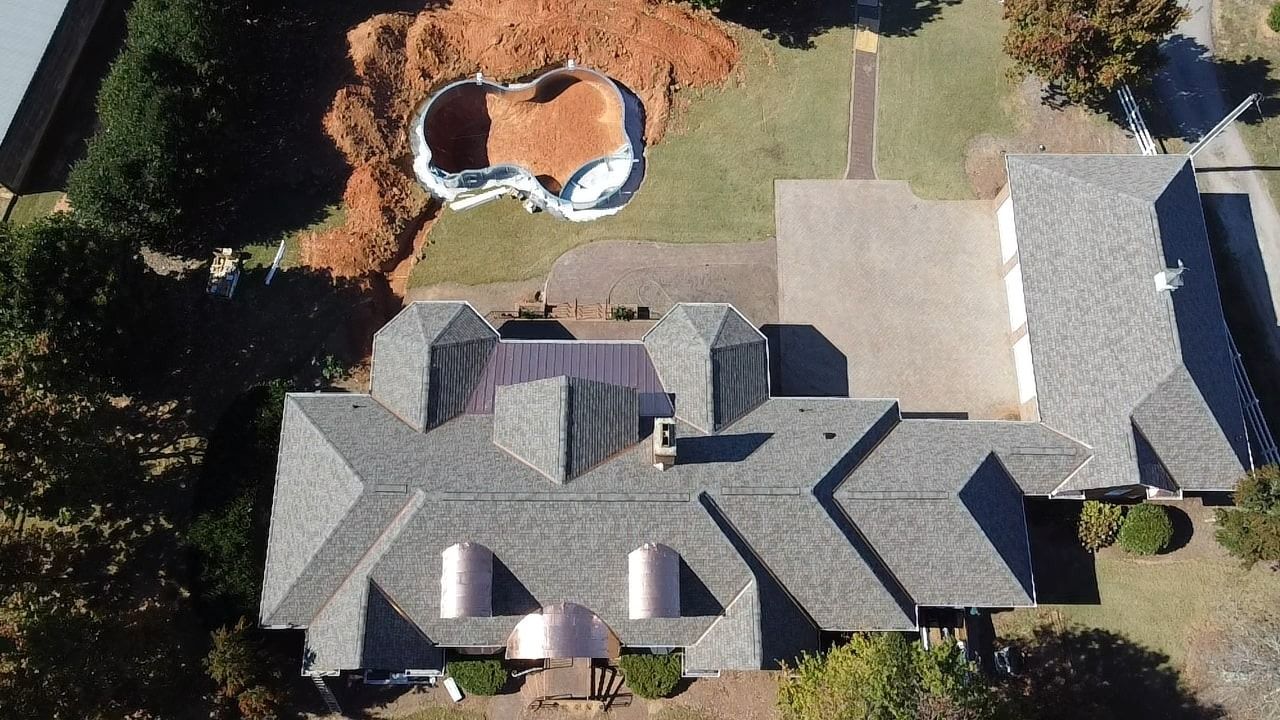 Aerial view of a large house with gray roofs, driveway, and backyard pool surrounded by trees.