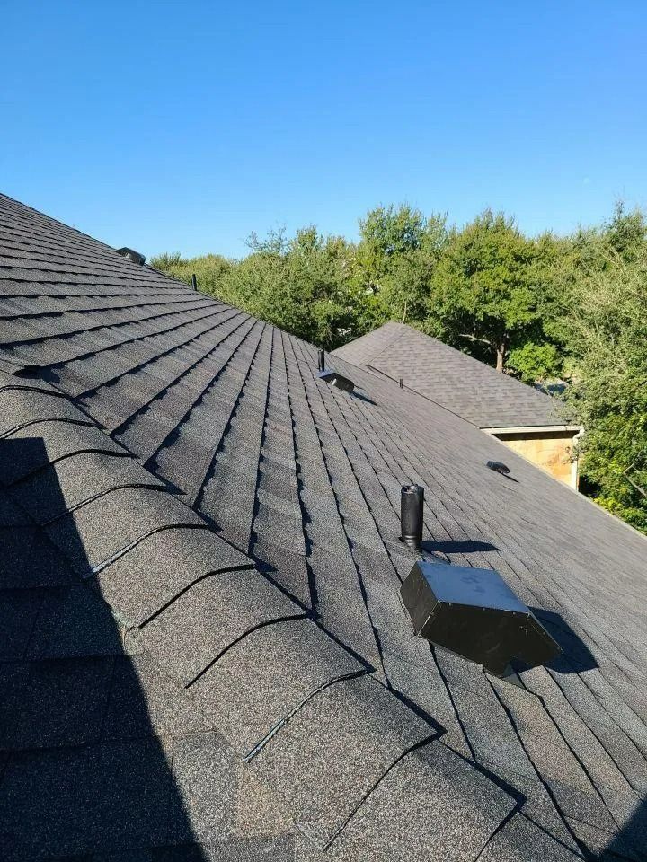 Black shingled roof with a skylight and vent under a clear blue sky, trees in the background