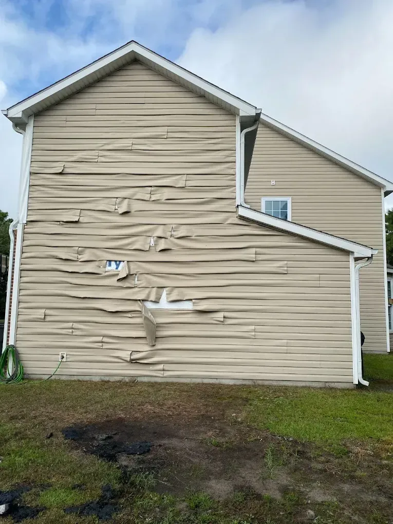 House siding severely dented and buckled on the side wall, with a damaged fence and muddy yard below.