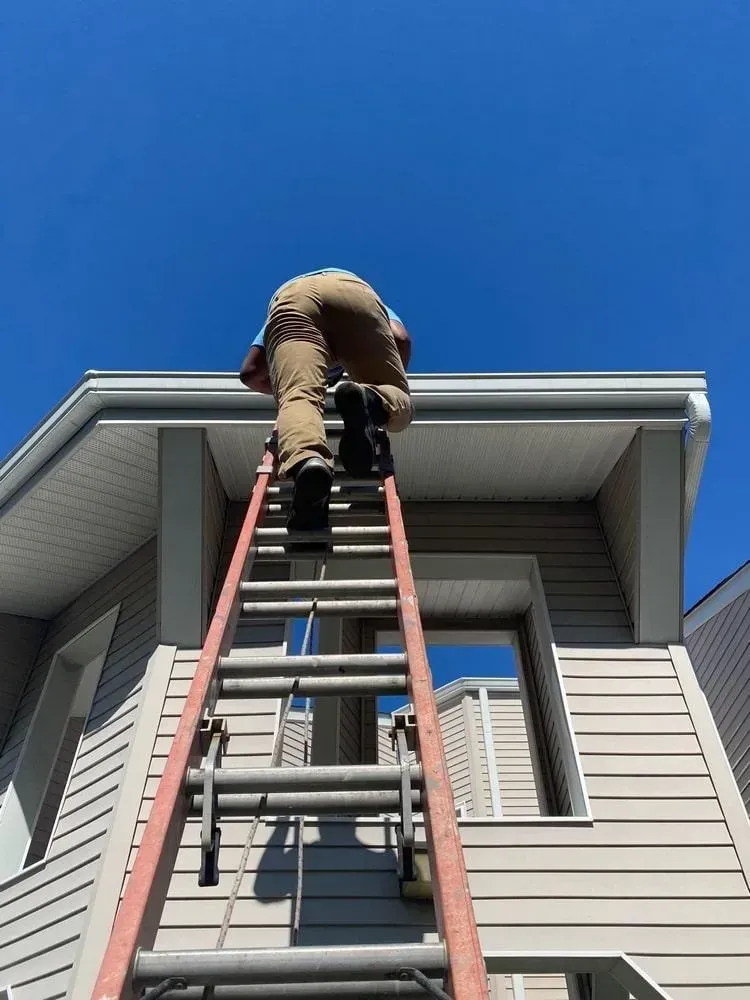 Worker on a ladder painting a house’s upper exterior beneath a blue sky