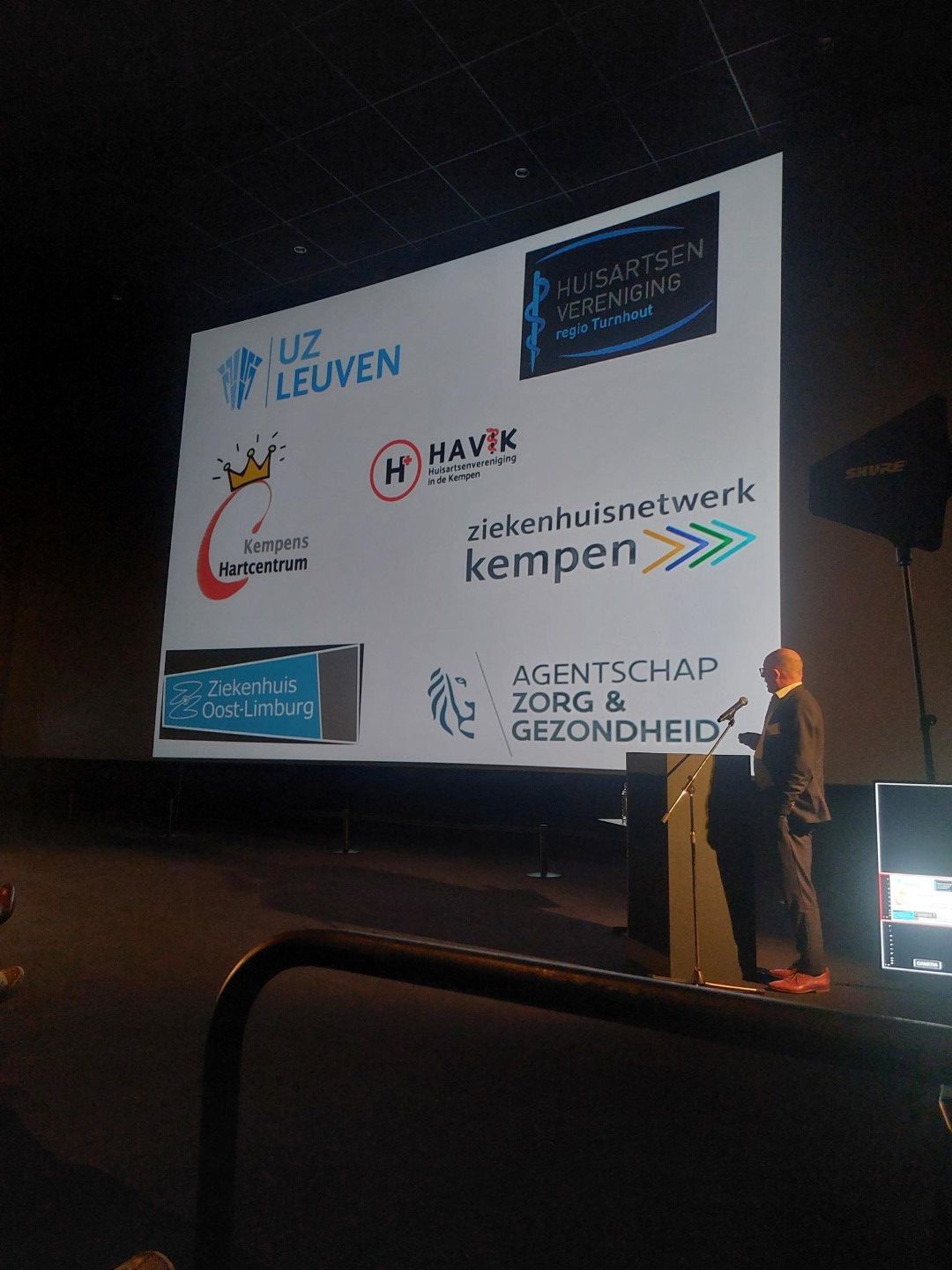 Een persoon spreekt op een podium naast een geprojecteerde dia met organisatielogo's in een donkere zaal.