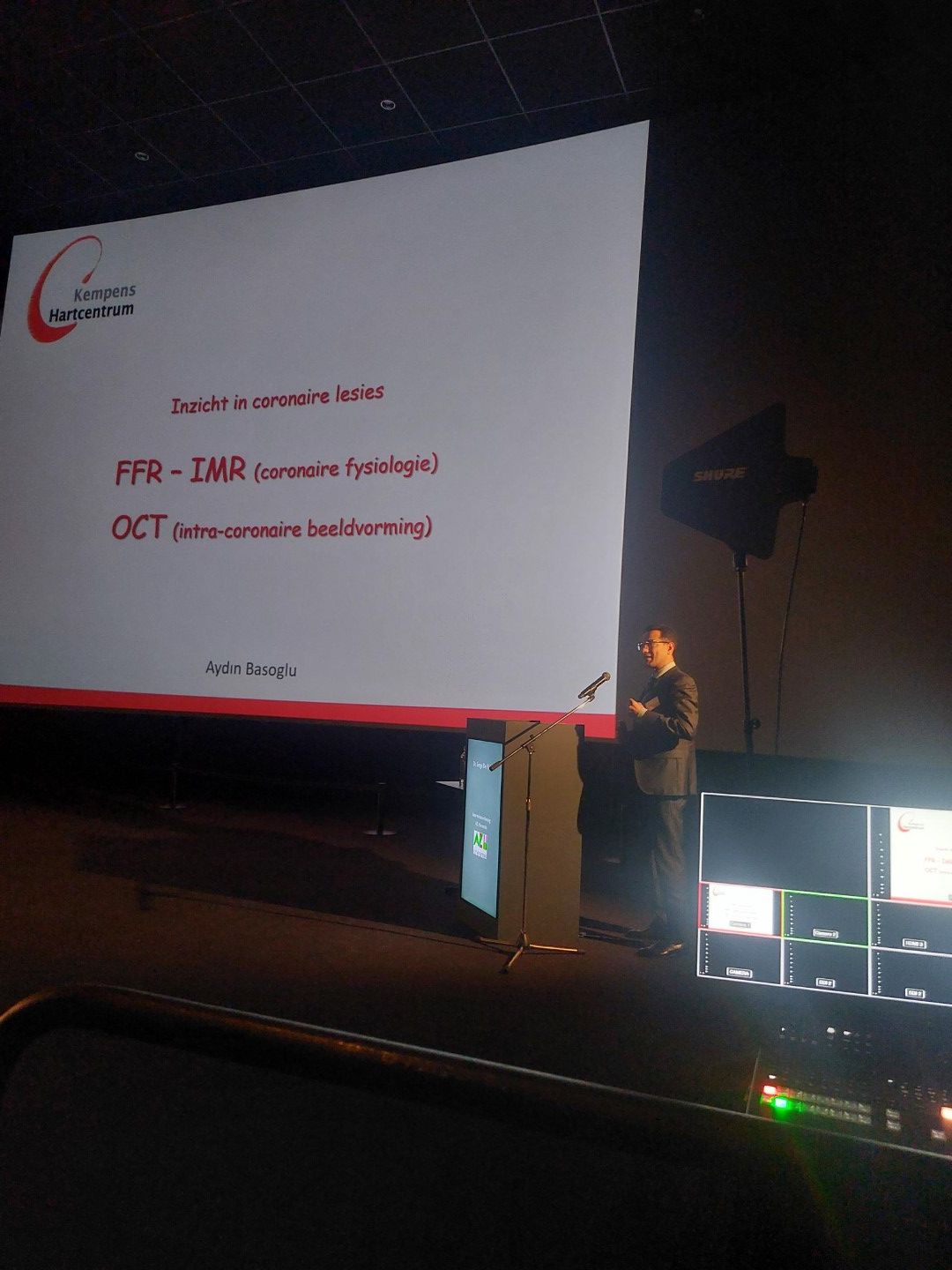 Een spreker op een conferentie staat naast een geprojecteerde dia over OCT en fundusfotografie in een donkere collegezaal.