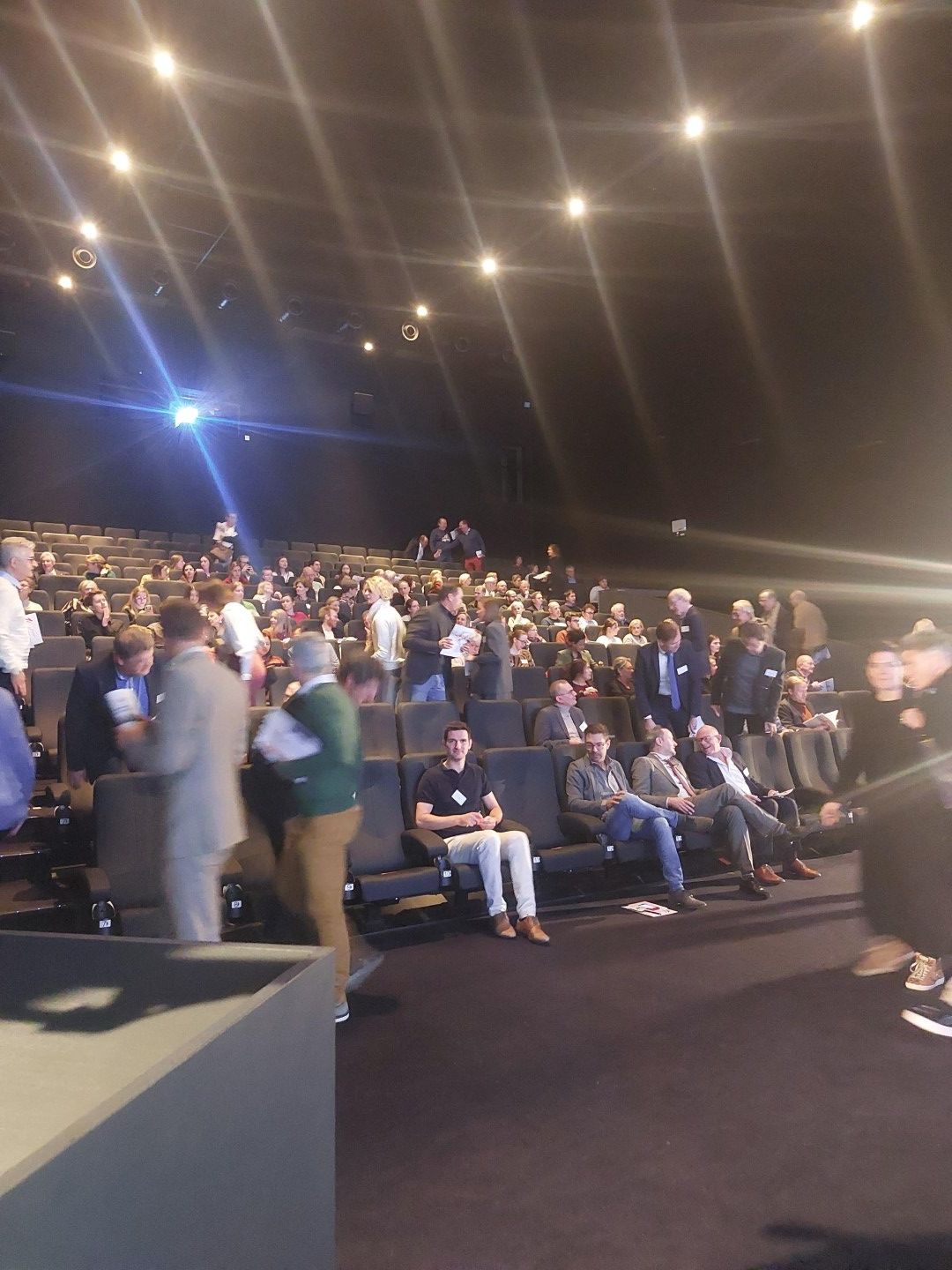 Het publiek zit in een donkere zaal onder fel plafondlicht en kijkt naar een evenement.