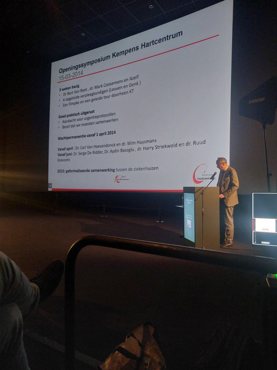 Spreker die een presentatie geeft vanaf een podium naast een grote geprojecteerde dia in een donkere zaal.