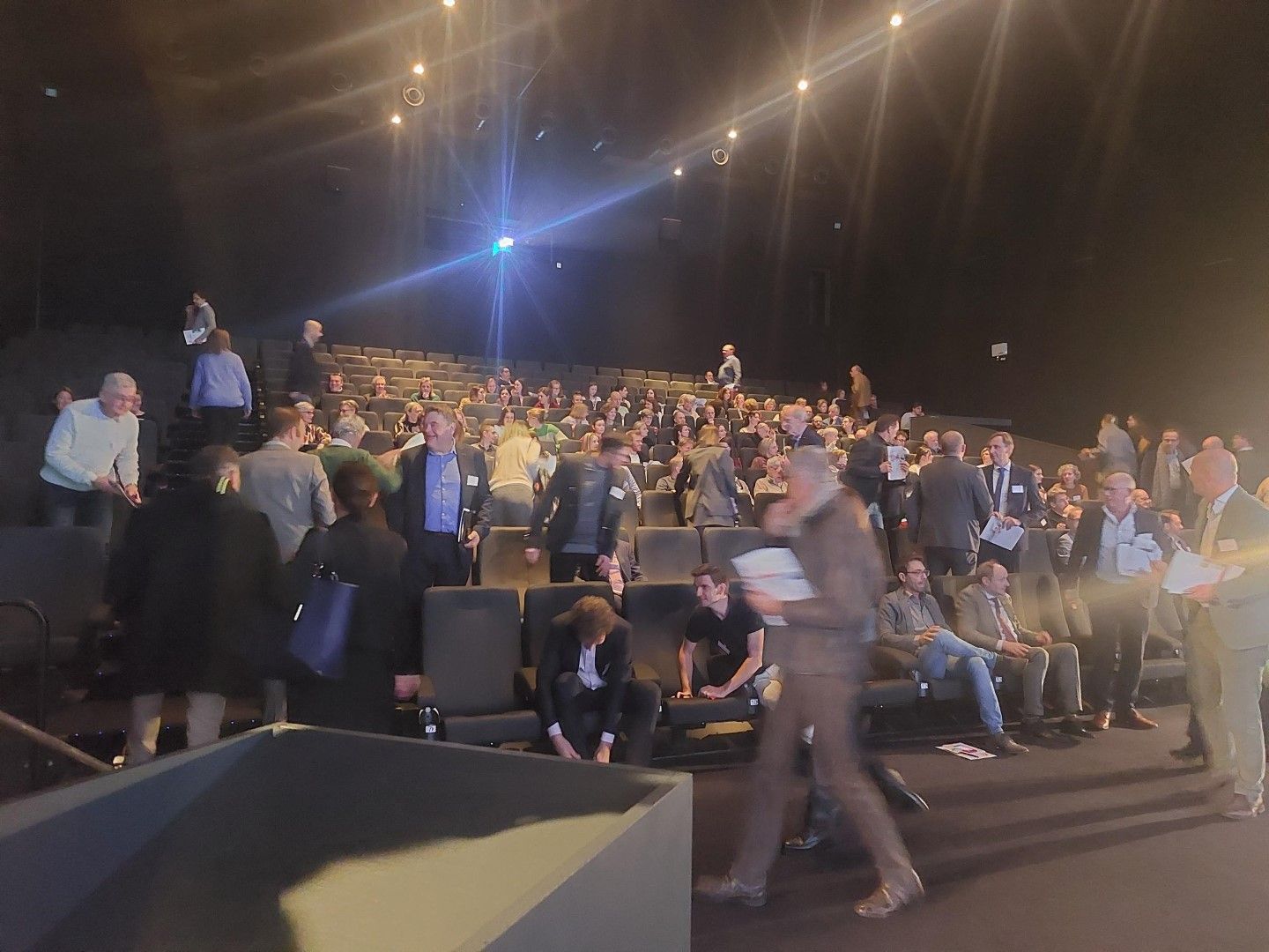 Een volle theaterzaal zit onder fel toneellicht, met een persoon die op de voorgrond loopt.