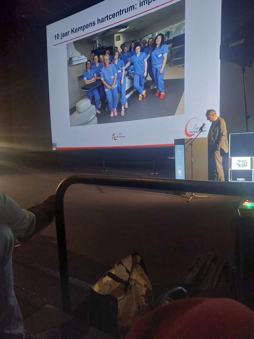 Spreker toont een groepsfoto op een groot scherm tijdens een conferentiepresentatie.